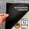 A hand holding up a corner of a black-backed floor mat, showing its texture and the German flag logo and a test rating. Náhled obrázku produktu
