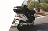 A person riding a white scooter on a road with trees and bushes in the background. Anteprima dell’immagine prodotto