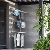 A white aluminum wall-mounted drying rack with three levels is shown on a balcony next to a brick wall, with laundry hanging on it. Green vines are growing on a dark pole to the right. Produktbild-Vorschau