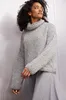 A woman wearing a gray turtleneck sweater and gray pants, posing against a ribbed light gray background. Produktbild-Vorschau