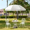 A white patio umbrella with fringe is set up over a white metal table and two chairs in a grassy garden area with palm trees and a building in the background. Produktbild-Vorschau