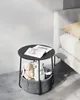 A round side table with a storage basket filled with stuffed toys and a book and glasses on top, placed next to a bed. Náhled obrázku produktu