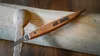 A carving knife and a partially carved wooden spoon are placed on a wooden surface. Anteprima dell’immagine prodotto