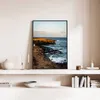 A framed picture of a coastal landscape is displayed on a shelf above other decorative items and books. Náhled obrázku produktu