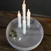 Four white candles are lit and placed in a round, light gray tray on a wooden surface. A small glass ornament with a sprig of pine is also in the tray. Produktbild-Vorschau