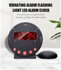 A black vibrating alarm clock with a red LED display sits on a light gray surface, with a green plant in the background. Náhľad obrázku produktu