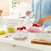 A person is placing cherry tomatoes into a clear plastic food storage container on a kitchen counter. Produktbild-Vorschau