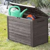 A garden storage box with its lid open, containing a green watering can and an orange and blue ball. The box is placed on a grassy lawn. Podgląd zdjęcia produktu