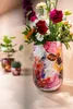 A close-up shot of a colorful vase filled with flowers, placed on a wooden surface. Two smaller potted plants are visible in the background. Produktbild-Vorschau