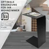 A black C-shaped side table placed in a modern living room, with a glass of white wine and a book on its surface. The background shows a blurred interior with stairs and furniture. Produktbild-Vorschau