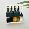 Four dark green glass bottles with gold spouts and a clear glass jar with a dark blue lid are arranged on a white shelf against a white wall. A blurred green leaf is visible in the upper right corner. Voir les images du produit