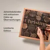 A person's hands are opening an advent calendar with a Christmas theme. The calendar is black with gold and copper decorative elements. Produktbild-Vorschau