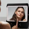 A woman is looking into a lighted makeup case mirror, touching the mirror with her finger. Produktbild-Vorschau