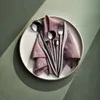 A set of silverware is neatly arranged on a pink napkin, which is placed on a white plate. The plate and silverware are set against a textured green background, with dramatic shadows cast by sunlight. Voir les images du produit