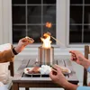 Two people are making s'mores over a small fire pit on a wooden table. Produktbild-Vorschau