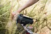 A person's hand is on the handlebars of a bicycle, with a phone mounted on a bag attached to the handlebars. The background is blurred tall grass. Anteprima dell’immagine prodotto