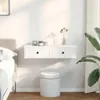 A white floating shelf with two drawers is mounted on a white wall above a white cylindrical stool. A bed with a beige headboard and white bedding is visible on the left. A vase with dried flowers and a large potted plant with green leaves are on the right Náhled obrázku produktu