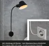 A black wall lamp with a flexible arm is mounted on a gray concrete wall. A close-up inset shows the lamp's power cord plugged into a white electrical outlet. Produktbild-Vorschau