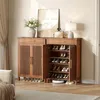 A wooden shoe cabinet with slatted doors and an open compartment displaying shoes, placed in a room with a sofa and curtains. Náhľad obrázku produktu