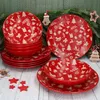 A set of red Christmas-themed dinnerware, including plates and bowls, is arranged on a wooden surface with a green, pine-needle-covered background. Decorative Christmas ornaments are scattered around the dinnerware. Produktbild-Vorschau