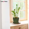 A green ceramic plant pot with a fish scale pattern sits on a wooden surface next to a window. Náhled obrázku produktu