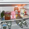The image shows the inside of a refrigerator with various food items and a pink pig-shaped object. Podgląd zdjęcia produktu