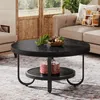 A round, black coffee table with a lower shelf is placed on a patterned rug in a living room setting. The table has a decorative bowl with pinecones and gold ornaments on top, and a red patterned box on the lower shelf. A sofa and an armchair are visible i Produktbild-Vorschau