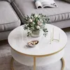 A round coffee table with a white top and gold accents, placed in a living room setting next to a gray sofa. Náhled obrázku produktu