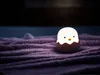 A small, glowing chick lamp in an eggshell sits on a textured purple blanket. Produktbild-Vorschau