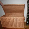 A hand is shown lifting the lid of a large wicker chest. A wine rack with bottles is visible in the background. Voir les images du produit
