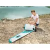 A woman is rolling up a paddleboard on a rocky shore next to a body of water. Náhľad obrázku produktu