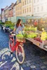 A woman is standing with her red bicycle at an outdoor market, with stalls of fresh produce in the background. Produktbild-Vorschau