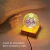 A hand is reaching out to touch a glowing crystal ball lamp on a wooden base, set on a soft surface. Produktbild-Vorschau