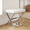 A white upholstered stool with a chrome X-frame base sits on a wooden floor next to a wall. A pair of white slippers are placed on the floor in front of the stool. A woven basket and a rug are visible in the background. Produktbild-Vorschau