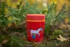 A red food jar with a cartoon illustration of a child holding a horse is placed on the ground amidst foliage and moss. Produktbild-Vorschau