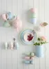 A collection of pastel-colored ceramic tableware, including cups, a teapot, egg cups, and a pitcher, arranged against a white paneled wall. Produktbild-Vorschau