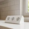 A white triple power strip sits on a speckled countertop in front of a tiled backsplash and a window. Produktbild-Vorschau
