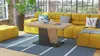 A modern living room with a yellow sectional sofa, a wooden coffee table, and a striped rug. Produktbild-Vorschau