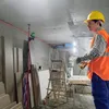 A construction worker in a yellow hard hat and orange safety vest is working indoors on a construction site. There are building materials and tools around him, including a laser level on a tripod. Náhled obrázku produktu
