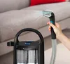 A hand holding a cleaning wand spraying water onto a couch, with a carpet cleaner in the foreground. Náhled obrázku produktu