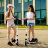 Two young girls are standing on a paved area with scooters in front of a modern building. Podgląd zdjęcia produktu