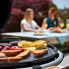 Two women are sitting at a table outdoors, with a charcoal grill in the foreground cooking food. Produktbild-Vorschau