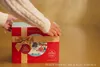 A person's hands are shown closing the lid of a red and gold advent calendar with a festive winter scene printed on it. Anteprima dell’immagine prodotto