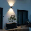 An outdoor wall light illuminates a white textured wall, a small tree, and a storage box on a paved patio at dusk. Produktbild-Vorschau