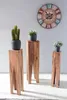 Three wooden plant stands of varying heights, each holding a potted plant, are arranged in front of a gray wall with a large decorative clock above. Náhled obrázku produktu