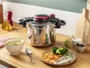 A pressure cooker is shown on a kitchen counter with a bowl of soup, a plate of food, and kitchen utensils. Podgląd zdjęcia produktu