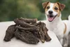 A pile of dried dog treats is placed on a table in front of a happy dog. Anteprima dell’immagine prodotto