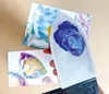 A hand holding an encaustic iron with blue paint on it, with two abstract art pieces in the background. Náhľad obrázku produktu