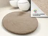 A round, fluffy beige bath mat is placed on a light-colored floor in front of two white toilets. Náhled obrázku produktu