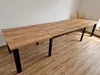 A long, extendable dining table with a wood-effect top and black metal legs, set against a plain wall and wooden floor. Produktbild-Vorschau